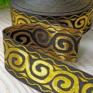 May include: A roll of black ribbon with a gold, swirling pattern. The ribbon is made of a woven material and has a metallic sheen. The design features a repeating motif of stylized curls and waves.