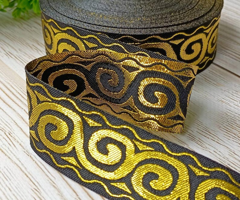 May include: A roll of black ribbon with a gold, swirling pattern. The ribbon is made of a woven material and has a metallic sheen. The design features a repeating motif of stylized curls and waves.