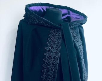 May include: A black hooded cloak with a purple satin lining. The cloak has detailed black floral lace trim along the hood and front edges. The cloak is displayed on a black mannequin head against a white backdrop.