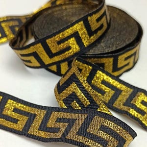 May include: Gold and black decorative ribbon with a Greek key pattern. The ribbon is woven with a metallic gold thread and a black background. Several rolls of the ribbon are visible, along with a length of the ribbon unrolled.