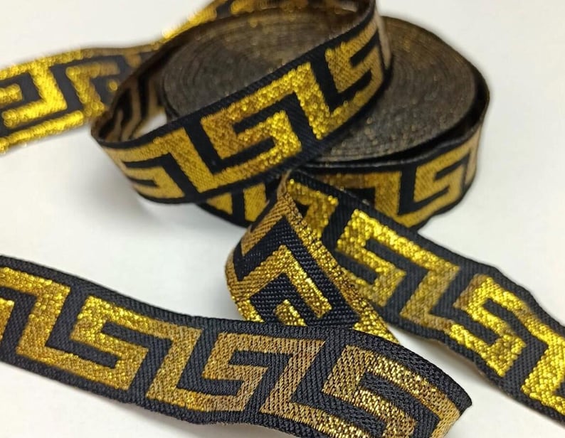 May include: Gold and black decorative ribbon with a Greek key pattern. The ribbon is woven with a metallic gold thread and a black background. Several rolls of the ribbon are visible, along with a length of the ribbon unrolled.