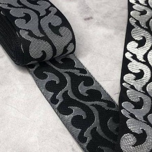 May include: Black and silver decorative ribbon with a scrolling, ornate pattern. The ribbon is rolled up, showing the edge and a section of the flat surface. The design features a repeating, stylized floral motif.
