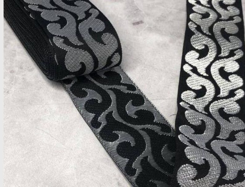 May include: Black and silver decorative ribbon with a scrolling, ornate pattern. The ribbon is rolled up, showing the edge and a section of the flat surface. The design features a repeating, stylized floral motif.