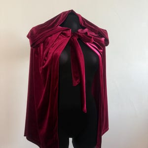 May include: A burgundy velvet cloak with a hood and a tie closure. The cloak is displayed on a black mannequin, showcasing its flowing design and rich color. The fabric has a soft sheen.
