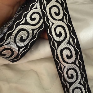 May include: Black ribbon with silver metallic swirls and a wavy border. The ribbon is held up against a white background. The ribbon is likely used for sewing, crafting, or embellishing garments.