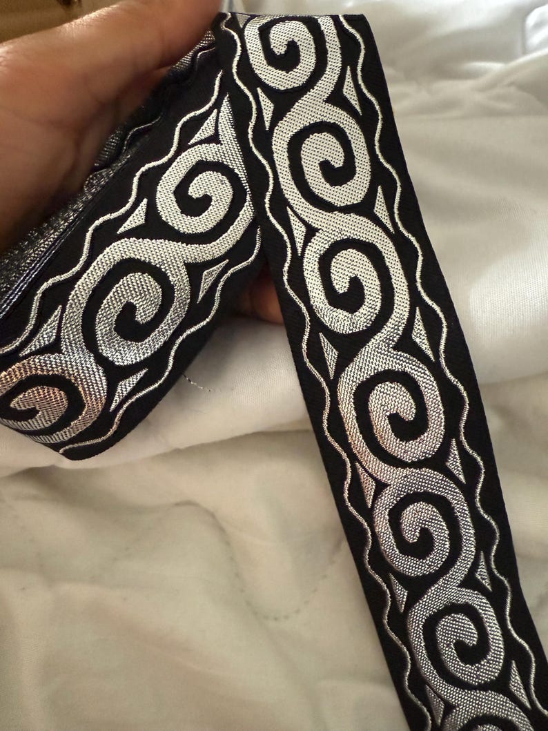 May include: Black ribbon with silver metallic swirls and a wavy border. The ribbon is held up against a white background. The ribbon is likely used for sewing, crafting, or embellishing garments.