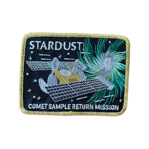 May include: A black and gold embroidered patch with the text "Stardust Comet Sample Return Mission". The patch features a stylized image of a spacecraft with a comet in the background.