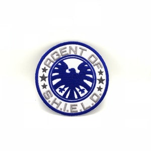 May include: A white and blue embroidered patch with the text "AGENT OF S.H.I.E.L.D." and a blue falcon logo in the center.