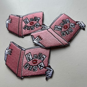 May include: Three pink embroidered patches shaped like open books. Each patch features the text "BURA BOOK" in white letters with a red lip in the center.