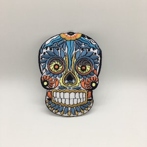May include: A colorful embroidered sugar skull patch with blue, yellow, orange, and white details. The skull is smiling with a detailed floral design.