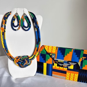 May include: A set of colorful African print fabric jewelry, including a necklace and earrings. The necklace is a thick rope of fabric with a knot at the center. The earrings are long and dangle. The fabric is a mix of blue, yellow, orange, and green.
