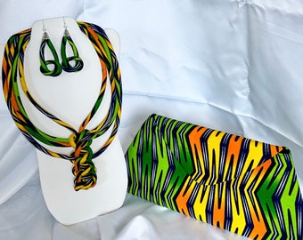 May include: A green, yellow, and orange patterned fabric necklace and matching clutch. The necklace is made of three strands of fabric twisted together and tied in a knot at the bottom. The clutch is a rectangular shape with a flap closure.