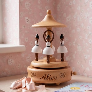 May include: A wooden music box carousel featuring three ballerina figures with dark skin tones, wearing white tutus. The base is light-colored wood, engraved with the name Alice and two bow icons. A pair of pale pink satin ballet slippers sits in front.
