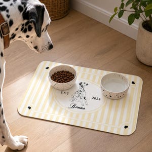 Puede incluir: Un tapete de alimentación rectangular para mascotas con un patrón de rayas verticales en amarillo claro y blanco. El centro presenta un marco ovalado blanco con una ilustración de dálmata, el texto EST 2024 y el nombre Bruno. Dos cuencos moteados.