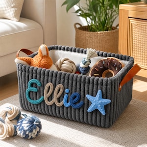 May include: A rectangular dark grey corduroy storage basket with tan leather handles. The front features the name Ellie in blue and beige yarn lettering, accompanied by a blue starfish appliqué. Inside are various plush toys and rope balls.