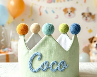 May include: A light green fabric crown featuring the name Coco in blue knitted lettering on the front. The top edge is decorated with five colorful yarn pom-poms in shades of orange, cream, light blue, lime green, and dark blue.
