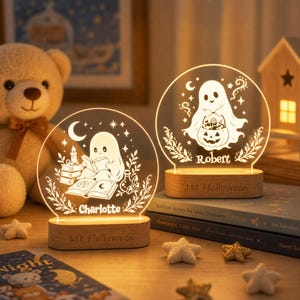 May include: Two circular acrylic night lights with wooden bases. One features a ghost reading a book with the name Charlotte, the other a ghost holding a pumpkin bucket with the name Robert. Both bases are engraved with 1st Halloween.