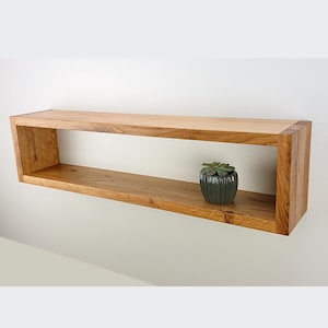 Può includere: Uno scaffale rettangolare in legno con un solo ripiano aperto. Lo scaffale è montato su una parete bianca e ha una piccola pianta verde in vaso sullo scaffale.