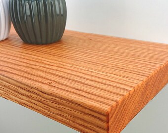 Könnte beinhalten: Eine Nahaufnahme eines Holzregals mit einer natürlichen Holzoptik. Das Regal ist aus massivem Holz gefertigt und hat eine glatte, polierte Oberfläche. Zwei kleine Topfpflanzen stehen auf dem Regal.
