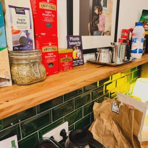 Pu&ograve; includere: Uno scaffale in legno con un paraschizzi in piastrelle verdi, che presenta diverse scatole di t&egrave;, un barattolo di vetro di erbe secche e una macchina per il caff&egrave;. Lo scaffale ha anche una cornice con una foto di due persone, una scatola di latte condensato e alcune caffettiere.
