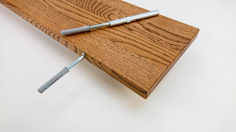 May include: A close-up of a wooden shelf with two gray plastic anchors and screws. The shelf is made of light brown wood with a natural grain pattern.