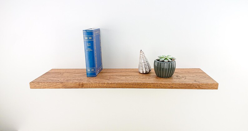 May include: A wooden shelf with a blue book, a small white ceramic cone, and a potted succulent plant. The shelf is made of natural wood with a light brown stain.
