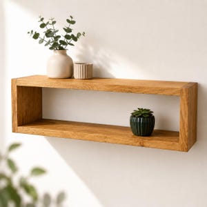 May include: A rectangular, open-front wooden shelf mounted on a white wall. The shelf holds a small beige vase with green sprigs, a ribbed beige container, and a small green potted plant. The wood has a natural, warm tone.