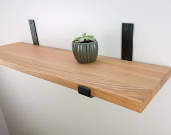 Könnte beinhalten: Ein schwebendes Holzregal mit natürlicher Oberfläche, das von schwarzen Metallhaltern getragen wird. Ein kleiner, gerippter, grauer Keramiktopf mit einer Sukkulentenpflanze steht auf dem Regal. Das Regal ist an einer weißen Wand befestigt.
