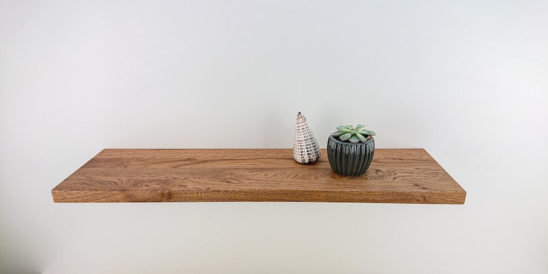 May include: A wooden shelf with a natural finish, featuring a small potted succulent plant and a white ceramic object. The shelf is mounted on a white wall.