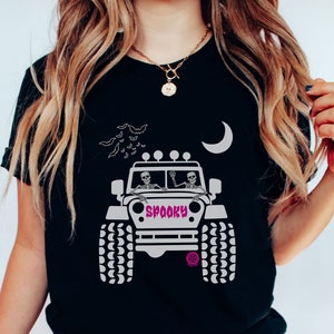 May include: Black t-shirt featuring a graphic of two skeletons driving a jeep with the word "Spooky" on the side. The jeep has a crescent moon and bats in the background.