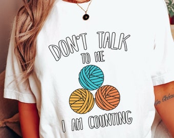 May include: White t-shirt with the text "Don't talk to me I am counting" and a graphic of three colorful balls of yarn.