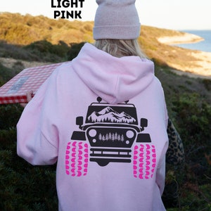May include: A light pink hoodie with a black graphic of a jeep with mountains in the background. The jeep has pink tires.