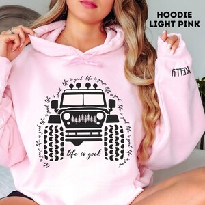 Puede incluir: Sudadera con capucha rosa claro con un gráfico negro de un jeep y la frase "Life is good" en un círculo. La manga muestra el nombre "KELLY". La prenda está hecha de tela suave.