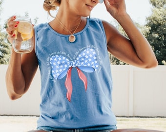 May include: A light blue tank top with a patriotic bow design featuring white stars, a blue bow, and red ribbons. The text "Flo Blue" is in the top right corner. The tank top is worn with denim shorts.