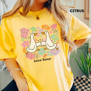 Könnte beinhalten: Ein gelbes T-Shirt mit dem Wort "Citrus" oben aufgedruckt. Das Shirt zeigt eine Grafik von zwei weißen Cartoon-Gänsen mit den Worten "Goose Bumps" darunter, umgeben von bunten Blumen.