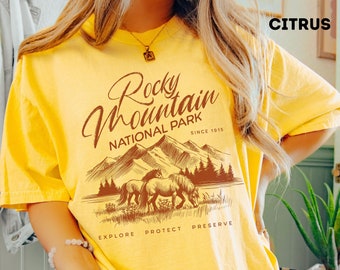May include: Yellow t-shirt with a brown graphic design that says "Rocky Mountain National Park" with a mountain range and two horses in the background. The text "Since 1915" is below the park name. The words "Explore, Protect, Preserve" are below the graphic.