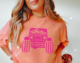 May include: A terracotta colored t-shirt with a pink graphic of a jeep with mountains in the background. The jeep has a front grill and tires with a tread pattern.