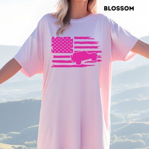 Könnte beinhalten: Ein rosafarbenes Oversize-T-Shirt mit einem abgenutzten amerikanischen Flaggen-Design, das eine rosafarbene Silhouette eines Jeeps zeigt. Der Text "BLOSSOM" befindet sich in der oberen rechten Ecke des Bildes.