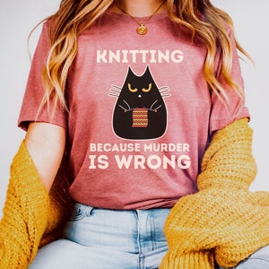 May include: A heather mauve t-shirt with a black cat knitting a striped scarf. The text on the shirt reads "Knitting because murder is wrong."