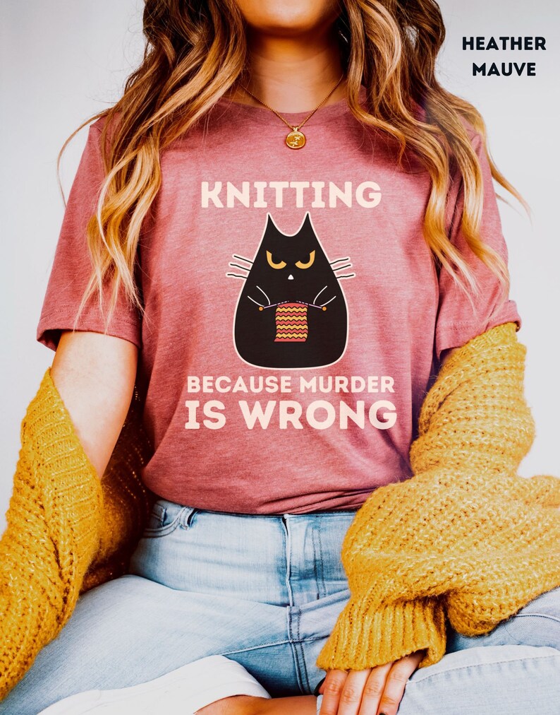May include: A heather mauve t-shirt with a black cat knitting a striped scarf. The text on the shirt reads "Knitting because murder is wrong."