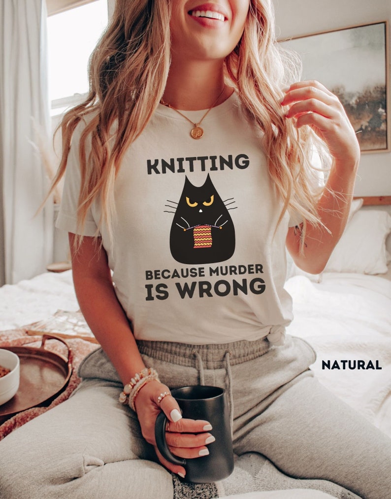 May include: White t-shirt with a black cat knitting a red and yellow scarf. The text on the shirt reads "Knitting because murder is wrong."
