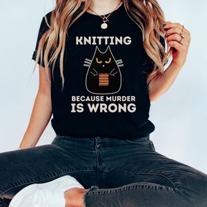 May include: Black t-shirt with a black cat knitting a red and yellow striped scarf. The text on the shirt reads "Knitting because murder is wrong."