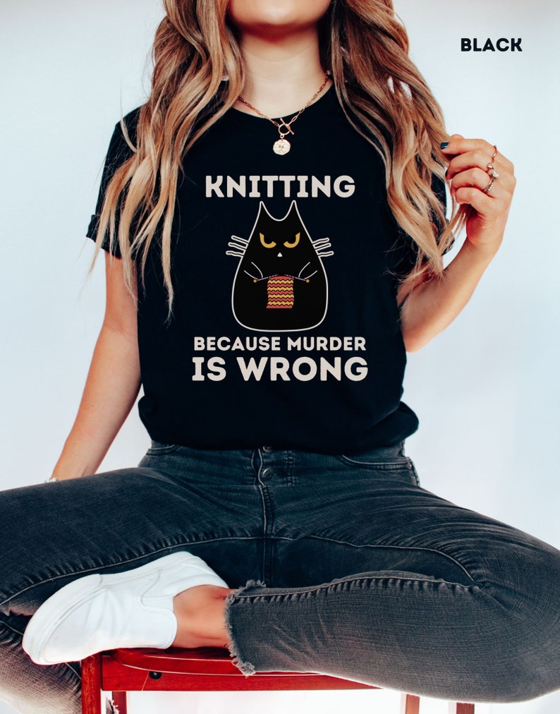 May include: Black t-shirt with a black cat knitting a red and yellow striped scarf. The text on the shirt reads "Knitting because murder is wrong."