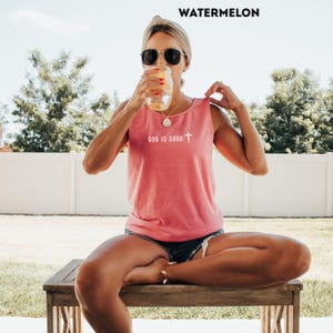 May include: A watermelon-colored tank top with the text "GOD IS GOOD" and a cross graphic. The top is sleeveless and made of a soft material. The word "WATERMELON" is printed at the top of the image.