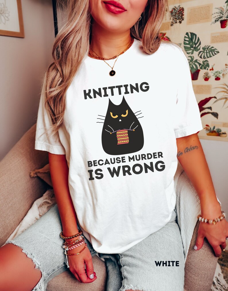 May include: White t-shirt with a black cat knitting a striped ball of yarn. The text on the shirt reads "Knitting because murder is wrong."