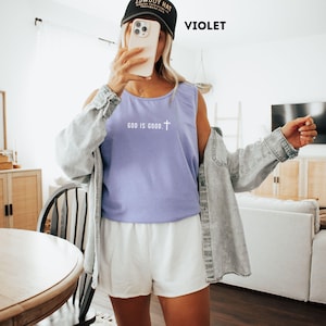 May include: A violet tank top with the text "GOD IS GOOD." and a cross graphic. The model is wearing a black baseball cap and a light blue denim jacket. The model is also wearing white shorts.
