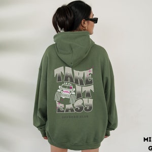 K&ouml;nnte beinhalten: Gr&uuml;ner Kapuzenpullover mit dem Text "Take It Easy" und einer Grafik eines Jeeps auf der R&uuml;ckseite. Der Text "Offroad Club" ist unter der Grafik gedruckt.