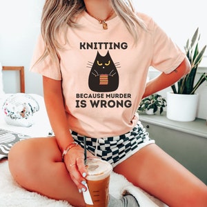 May include: A woman wearing a heather peach t-shirt with the text "Knitting because murder is wrong" and a black cat illustration holding knitting needles. The cat has yellow eyes and is knitting a striped scarf.