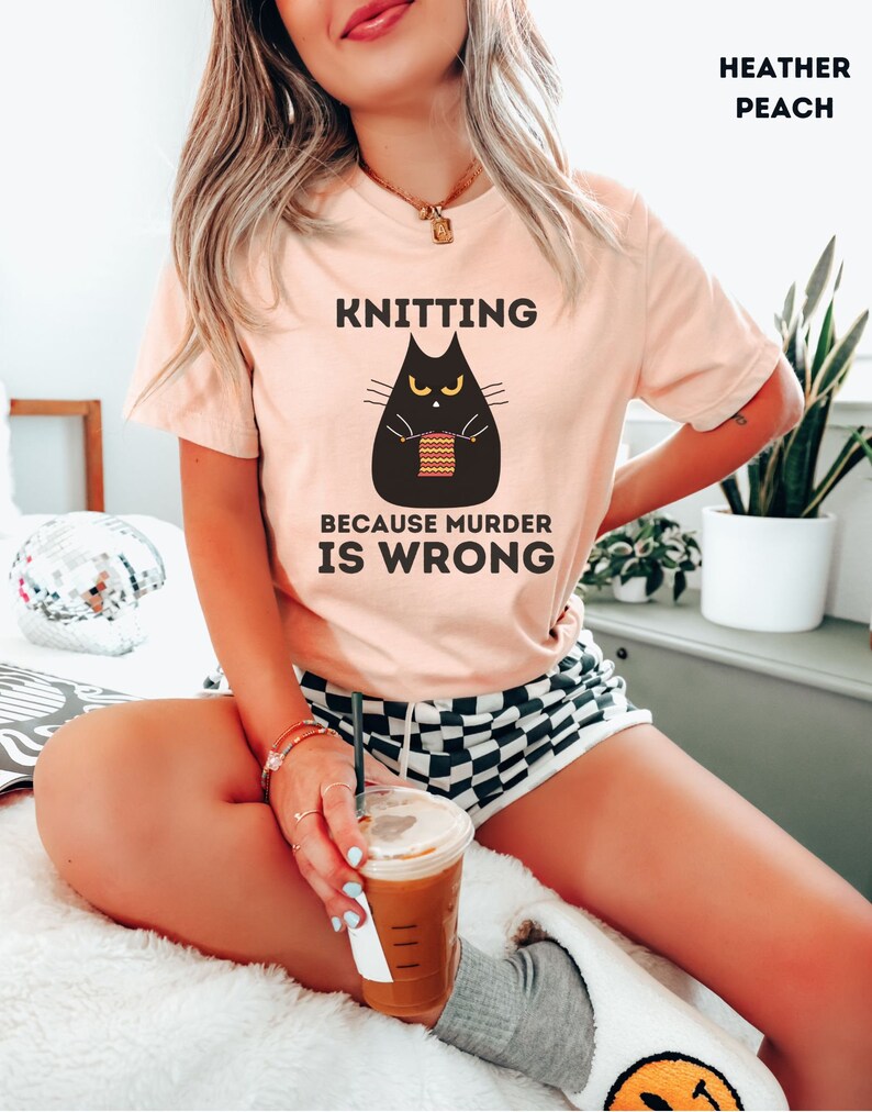 May include: A woman wearing a heather peach t-shirt with the text "Knitting because murder is wrong" and a black cat illustration holding knitting needles. The cat has yellow eyes and is knitting a striped scarf.