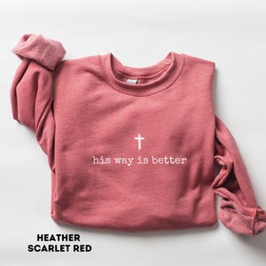 May include: A heather scarlet red sweatshirt with the text "his way is better" and a white cross. The long-sleeved crew neck top is folded on a white surface. The words "HEATHER SCARLET RED" are printed at the bottom.
